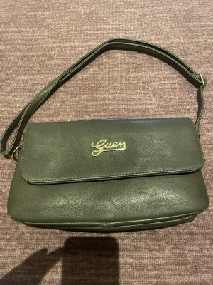 Olivgrön/grå axelremsväska från Guess - Stilren olivgrön/grå handväska från Guess i mjukt konstläder. Väskan har ett guldfärgat Guess-emblem på locket och justerbar axelrem. Klassisk rektangulär form och enkel design som passar till många outfits. Lite sminkfläckar inuti men inget som stör😊