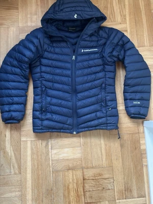 Mörkblå dunjacka från Peak Performance - Snygg mörkblå dunjacka från Peak Performance med huva och dragkedja framtill. Jackan har två sidofickor med dragkedja och diskret logga på bröstet samt R&D-detalj på ärmen. Perfekt för kalla vinterdagar och riktigt lätt att bära.