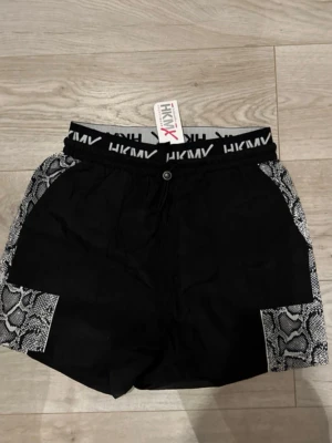 Svarta shorts med ormmönster från Hunkemöller - Säljer ett par svarta shorts från hunkemöller med elastisk midja och logotyp. Sidorna och fickdetaljerna har grått ormmönster som ger en cool kontrast. Perfekta för gymmet eller streetwear. Materialet känns lätt och bekvämt.
