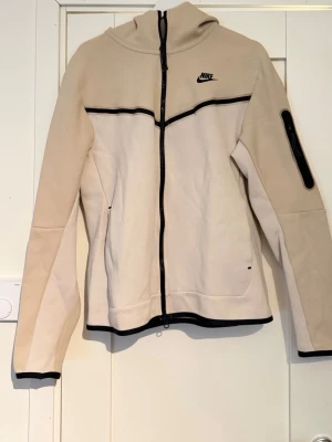 Beige hoodie från Nike med dragkedja - Snygg beige hoodie från Nike med hel dragkedja framtill och svarta detaljer. Hoodien har en huva, svarta linjer över bröstet och ärmslut samt en praktisk ficka med dragkedja på ena ärmen. Tillverkad i mjukt bomullsmaterial för skön känsla.