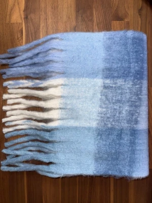 Rutig halsduk i bått och vitt - Mjuk och fluffig halsduk med rutigt mönster i blått och vitt. Halsduken har långa fransar i båda ändar och är perfekt för att hålla värmen under kalla dagar. Materialet känns ulligt och ger en mysig känsla. Halsduken liknar ”Acne Studios halsdukarna och är oanvänd (ny).