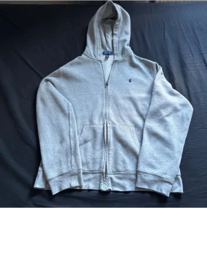 Grå zip-hoodie från Polo Ralph Lauren - Klassisk grå hoodie med dragkedja från Polo Ralph Lauren. Har huva, känguruficka och liten broderad logga på bröstet. Perfekt för en chill och avslappnad stil. Storlek Barn Xl. Passar ca 165-170. Mudden är lite skrynklig men går fixa lätt med strykjärn
