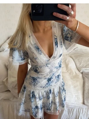 Blommig vit och blå kortklänning - Säljer denna sååå perfekta klänningen i så bra kvalitet. Superbra skick och så smickrande och  fin passform💕 Se mått! Midja rakt över: ca 38cm, höfter rakt över: ca 45, byst rakt över: ca39cm, längd: ca 83cm.