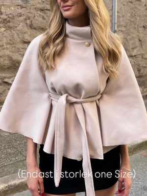 Beige cape-kappa med knytband, finns även i svart! - Stilren beige cape-kappa med hög krage och knytband i midjan. Kappan har vida ärmar och en dekorativ knapp vid halsen. Endast i one size. Finns även i svart