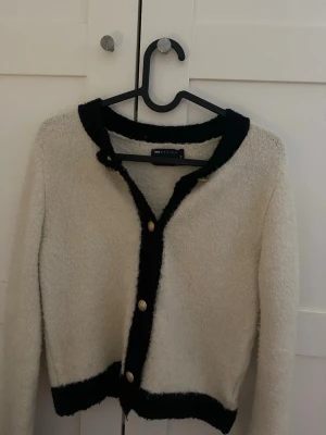 Krämvit kofta med svarta detaljer ASOS - Säljer en krämvit kofta från ASOS DESIGN i storlek XS. Koftan har svarta kanter och guldknappar framtill, samt en mjuk, fluffig textur. Långärmad och kortare modell som ger en trendig look.