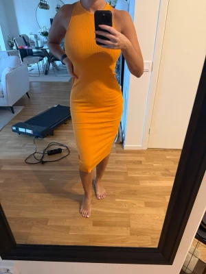 Gul ribbad midiklänning med slits - Säljer en snygg gul ribbad klänning med halterneck och korsade band i ryggen. Klänningen är figurnära, ärmlös och har en slits bak för extra rörelsefrihet. Perfekt för dig som vill sticka ut med färg och stil i sommar!