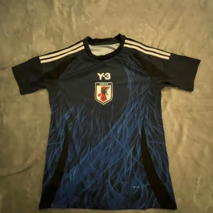 Japan Y-3 fotbollströja blå - Snygg blå fotbollströja från Y-3 med Japan JFA-emblem på bröstet. Tröjan har svarta och vita detaljer, klassiska Adidas-ränder på axlarna och ett coolt blått mönster framtill. Perfekt för dig som gillar sportig stil och fotboll.