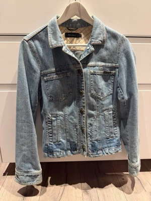 Ljusblå jeansjacka Prêt à Porter - Snygg ljusblå jeansjacka från Prêt à Porter med klassisk krage, fyra fickor framtill och coola metallknappar med logga. Rutigt innerfoder ger extra stilpoäng. Perfekt att slänga över en hoodie eller t-shirt för en avslappnad look.