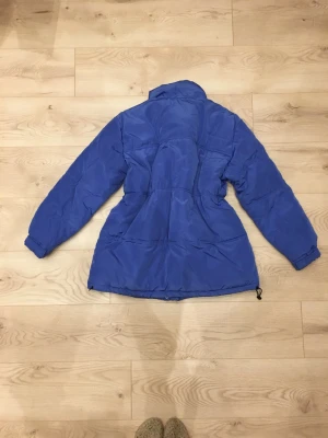 Blå puffjacka med hög krage - Snygg blå puffjacka i lite längre modell och med hög krage. Jackan har två framfickor, stängs med dragkedja och knappar fram, och har elastiska muddar vid ärmsluten.