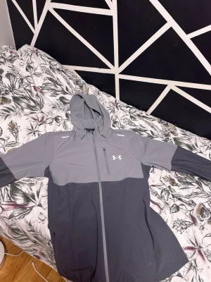 Grå vindjacka från Under Armour, Storlek M - Snygg tvåtonad grå vindjacka från Under Armour med huva och hel dragkedja framtill. Jackan har en bröstficka med dragkedja och diskret logga på vänster sida. Tillverkad i lätt och vindtätt material, perfekt för blåsiga dagar. Sportig look med kontrastpaneler på ärmar och kropp.