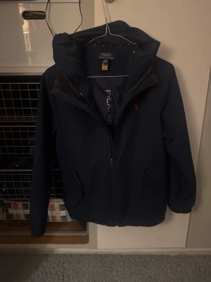 Mörkblå vindjacka från Polo Ralph Lauren - Snygg mörkblå vindjacka från Polo Ralph Lauren med huva och dragkedja framtill. Jackan har två sidofickor och en liten röd logga på bröstet. Perfekt för blåsiga dagar och enkel att matcha med olika outfits. Har ett litet brännmärke längst in på axeln som man inte ser när man använder den. Kostar 1600-1900 ny