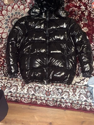 Svart glansig jacka med huva - Säljer en svart, glansig jacka med huva och dragkedja framtill. Jackan har två stora fickor med dragkedja och elastiska muddar vid ärmsluten. Perfekt för kalla vinterdagar och riktigt snygg streetstil. 