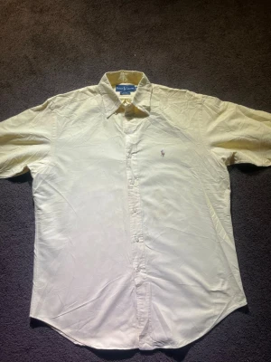  Gul skjorta från Ralph Lauren M - Säljer en klassisk gul skjorta från Ralph Lauren i storlek M. Skjortan är tillverkad i 100% bomull och har knappar framtill samt den ikoniska broderade loggan på bröstet. Perfekt för dig som gillar stilrena och tidlösa plagg.