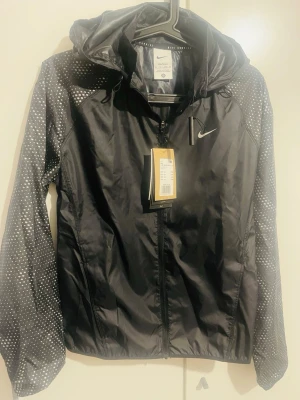 Svart Nike vindjacka med prickiga ärmar - Nike vindjacka i svart med huva och hel dragkedja. Ärmarna har ett coolt vitt prickigt mönster som ger jackan extra stil. Lätt och smidig polyester, perfekt för utomhusaktiviteter. Swoosh-logga på bröstet och justerbar huva.