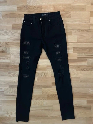 Svarta skinny jeans från Amiri - Svarta jeans från Amiri med skinny passform och snygga slitningar på benen. Bakom slitningarna finns coola detaljer med små silverfärgade nitar som ger extra edge. Klassisk femficksmodell med dragkedja och knapp framtill.
