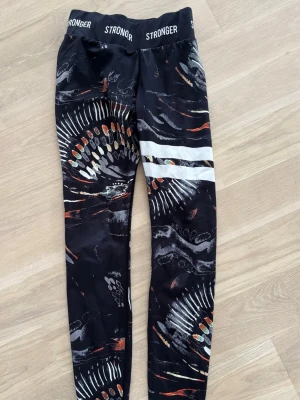 Svarta leggings från Stronger med mönster - Snygga svarta leggings från Stronger med coolt abstrakt mönster i vitt, orange och grått. De har bred resår i midjan med logga och två vita ränder på ena benet. Perfekta för träning eller chill. Materialet är stretchigt och känns skönt mot huden.