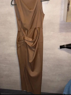 Brun långklänning med drapering - Snygg brun långklänning med ärmlös design och cool drapering framtill som ger en unik look. Klänningen har en slits nedtill och är gjord i ett slätt, glansigt material som känns lyxigt. Perfekt för dig som vill sticka ut med stilren elegans.