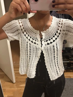Vit virkad kofta med kort ärm - Säljer en superfin vit virkad kofta med korta ärmar och dekorativa hålmönster. Koftan har två knappar framtill och vågig kant längs halsringningen och nederkanten. 