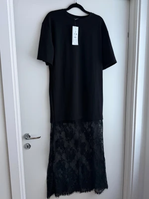 NY slutsåld populär tshirttklänning med spets från  Gina Tricot - Säljer en svart långklänning från Gina Tricot i t-shirtmodell med korta ärmar och rund hals. Nederdelen är i transparent spets med blommigt mönster som ger en snygg kontrast till den enkla överdelen. Perfekt för dig som vill ha en edgy och trendig look.