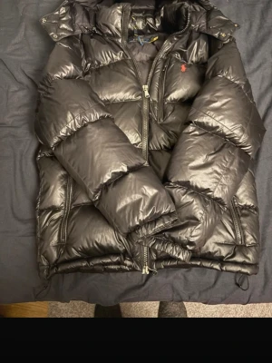 Svart pufferjacka från Polo Ralph Lauren - Svart pufferjacka från Polo Ralph Lauren med glansig finish och klassisk röd logga på bröstet. Jackan har huva, flera dragkedjor och är quiltad för extra värme. Materialet är polyester och dun, perfekt för kalla dagar. Snygg och stilren design som sticker ut. Tveka inte att hör av dig.(pris ej hugget i sten)