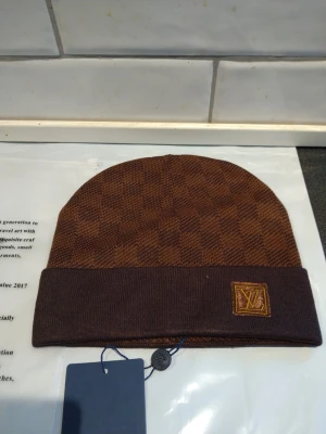 Brun rutig Louis Vuitton mössa - Snygg brun mössa från Louis Vuitton med klassiskt rutigt mönster och uppvikt kant. Mössan har en gul LV-logga framtill och är tillverkad i ett mjukt stickat material som känns skönt mot huvudet. Perfekt för dig som vill ha en lyxig accessoar till hösten och vintern.
