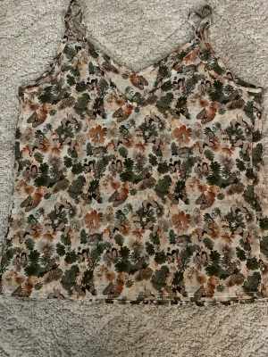 Blommig linnetopp med fjärilar - Säljer en söt linnetopp med justerbara axelband och blommigt mönster i grönt, beige och orange. Toppen har även fjärilar i printet och är gjord i ett lätt, syntetiskt material som känns luftigt. Perfekt för varma dagar och har en avslappnad passform.