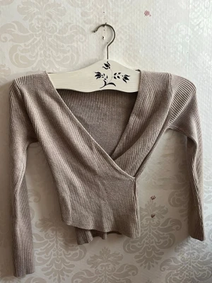 Beige omlottopp från SHEIN - Säljer en beige omlottopp från SHEIN i storlek XS. Toppen är ribbad med lång ärm och har en snygg v-ringad omlottdesign framtill. Materialet är mjukt och stretchigt, perfekt för att skapa en chill och stilren look.