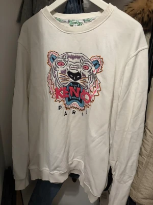 Vit sweatshirt med tiger från Kenzo - Säljer en vit sweatshirt från Kenzo med stort broderat tigerhuvud i flera färger och logga på bröstet. Tröjan har rund hals, ribbade muddar och lång ärm. Perfekt statement-plagg för dig som gillar coola prints och streetstyle.