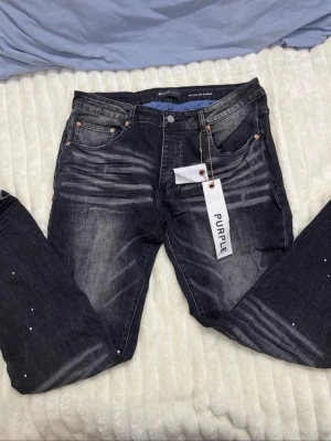 Svarta skinny jeans från Purple Brand - Säljer ett par svarta skinny jeans från Purple Brand med coola slitningar och ljusa detaljer på benen. Jeansen har låg midja och klassisk femficksdesign med metallnitar. Materialet är jeans och passformen är slim med smal benvidd. Perfekt för dig som gillar streetwear.