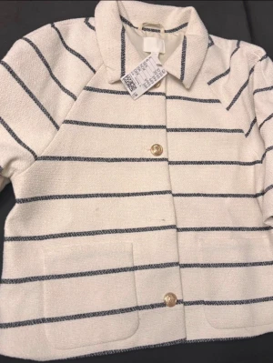 Vit och marinblå randig kavaj från H&M - Snygg kavaj från H&M i vit med marinblå ränder. Modellen har klassisk krage, två stora fickor framtill och guldiga knappar. Materialet är strukturerat och känns lyxigt. Perfekt för dig som gillar stilrena och trendiga plagg.