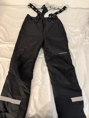 Svarta täckbyxor från Cross Sportswear - Svarta täckbyxor från Cross Sportswear med justerbara hängslen och kardborreband i midjan så att man kan justera storleken runt midjan. Byxorna har dragkedja och knappstängning, två fickor fram och reflexdetaljer vid bensluten. Perfekta för vintern och utomhusaktiviteter. 