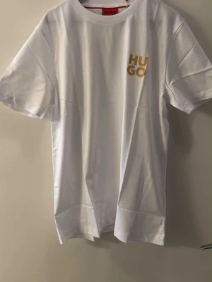 Vit t-shirt från Hugo med gul logga - Vit t-shirt från Hugo med rund hals och korta ärmar. På bröstet finns en gul tryckt HUGO-logga som ger en clean och modern vibe. Materialet är mjuk bomull som känns skönt mot huden. Perfekt för dig som gillar stilrena och enkla plagg med en twist.