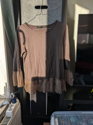 Brun blus med volang från Prêt à Porter - Säljer en brun långärmad blus från Prêt à Porter i storlek M. Toppen har transparenta volangdetaljer vid ärmslut och nederkant som ger en cool och unik look. Rund halsringning och loose passform, perfekt att styla med jeans eller kjol.