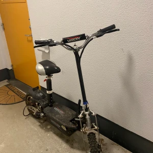 Svart el-scooter med sadel från Viron - Elektrisk scooter från Viron i svart med bred sadel i svart och vitt. Scootern har grova däck, fjädring fram och bak samt styre med digital display. Passar för både stadskörning och enklare terräng. Batteriet behövs bytas men har aldrig orkat göra det. Den går i 25-30km/h och det som behövs fixa är: Bromsar, lufta däcken. Buda på !