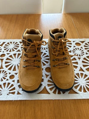 Timberland kängor i mocka, strl 43 - Säljer ett par snygga Timberland kängor i beige mocka med svarta detaljer och snörning. Skorna har rund tå, platt sula och är tillverkade i mocka och textil. Perfekt för dig som gillar outdoor-vibe och vill ha något slitstarkt och stilrent.