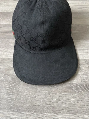 Svart Gucci keps  - Svart keps från Gucci med diskret GG-monogram över hela ytan. Klassisk böjd skärm och ikoniska gröna och röda ränder på sidan. Tillverkad i polyester och bomull, med etikett som visar 'Made in Italy'. Snygg och lyxig accessoar för dig som gillar designer.