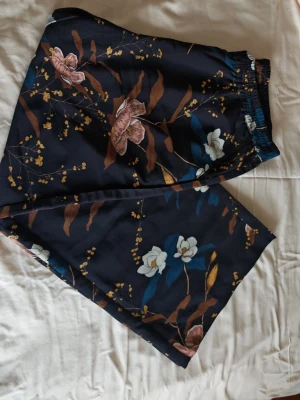 Irene Blommiga byxor från Honey, storlek L - Säljer ett par svarta byxor från Honey med blommigt mönster i vitt, rosa och brunt. Byxorna har elastisk midja och är gjorda i ett mjukt, lätt material som känns skönt mot huden.100% polyster. Inner mått 76. Perfekta för dig som gillar avslappnad och färgstark stil.