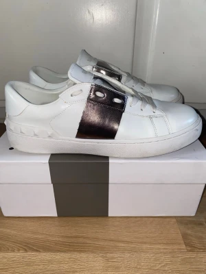 Valentino Garavani sneakers med metallic - Snygga vita sneakers från Valentino Garavani med en bred metallic-silver rem över sidan och coola nitar på hälen. Skorna har snörning och en platt sula, tillverkade i skinn för en lyxig känsla. Perfekt för dig som gillar statement-detaljer och exklusiv design.