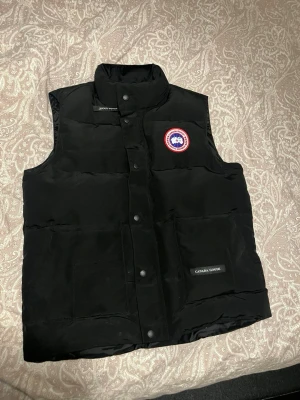 Svart dunväst från Canada Goose - Svart dunväst från Canada Goose med hög krage och tryckknappar framtill. Västen har ett klassiskt Canada Goose-märke på bröstet och praktiska fickor med logotyp. Perfekt för kyliga dagar och har en stilren, quiltad design.