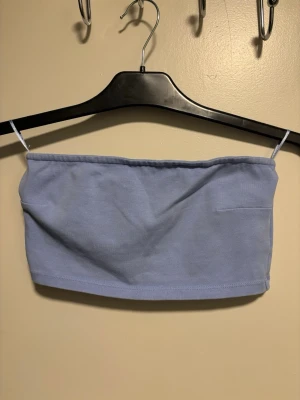 Ljusblå bandeau tubtopp - Säljer en enkel och stilren ljusblå bandeau tubtopp. Toppen är ärmlös och axelbandslös, med en rak och croppad passform. Materialet känns mjukt och stretchigt, troligtvis bomull eller syntet. Perfekt för varma dagar och att styla med jeans eller kjol.