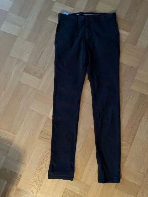 Svarta chinos från Tommy Hilfiger - Snygga svarta chinos från Tommy Hilfiger med klassisk rak passform och diskret logga ovanför bakfickan. Byxorna har bälteshällor, sidofickor och bakfickor med knapp. Perfekta för en clean och stilren look.