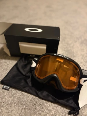 Oakley skidglasögon - Bilderna visar ett par Oakley skidglasögon med svart ram och orange lins, samt tillhörande förvaringspåse och originalkartong.                     Skick: 10/10 helt nya                                               Frakt: Inom 24h                                                        Hör gärna av dig med fler frågor eller funderingar!