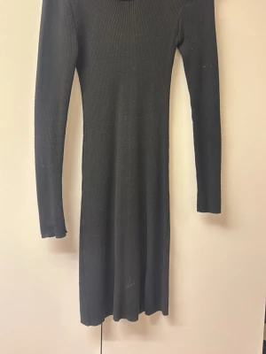 Svart stickad långärmad klänning - Säljer en svart stickad klänning med lång ärm och ribbad struktur. Klänningen har en enkel, figurnära passform och är knälång. Perfekt att styla med smycken eller boots för en cool look.