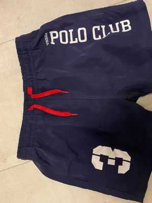 Vinson Polo Club badbyxor - Hej! Säljer snygga marinblå badbyxor från Vinson Polo Club med vit logotyp och text framtill. De har röd snörning i midjan och meshfoder på insidan för extra komfort. Perfekta för strand eller poolhäng, med elastisk midja och normal passform.