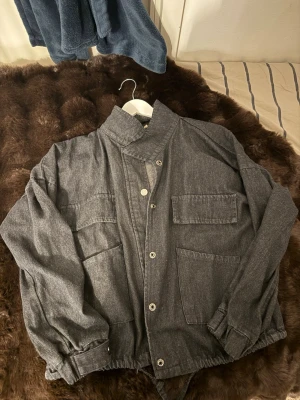 Grå svart denim jacka med knappar - Säljer en grå  svart overshirt med hög krage och fyra stora fickor framtill. Jackan har knappar längs hela framsidan och elastiska muddar vid ärmsluten. Materialet känns som bomull och ger en avslappnad vibe. Perfekt att slänga över en hoodie eller t-shirt. Den har även en kortärmad hoodie som man kan under, går att köpa med denna! Pris kan diskuteras! 