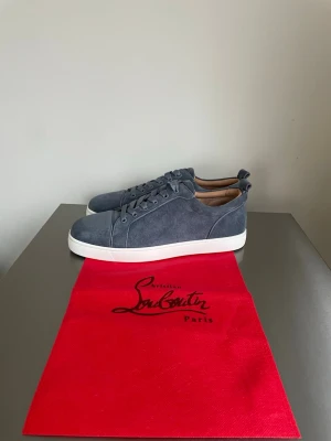Grå mocka sneakers från Louboutin - Snygga grå sneakers från Christian Louboutin i mjuk mocka med vit platt sula och klassisk röd undersida med logotyp. Skorna har rund tå, snörning och beige innersula med guldfärgad branding. Perfekt för dig som gillar exklusiv stil och detaljer.