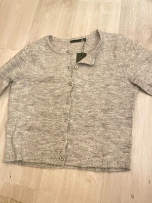 Ljusgrå kofta från 157, storlek S - Mysig ljusgrå kofta från 157 i storlek S. Koftan har rund halsringning, långa ärmar och knappar hela vägen framtill. Perfekt att slänga över en t-shirt eller linne för extra lager. Enkel och stilren design som passar till det mesta.