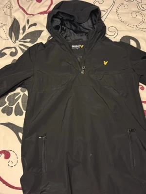 Svart vindjacka från Lyle & Scott - Svart vindjacka från Lyle & Scott med huva och halv dragkedja framtill. Jackan har två fickor med dragkedja och den klassiska gula loggan på bröstet. Tillverkad i lätt syntetmaterial, perfekt för blåsiga dagar. Enkel och stilren design.
