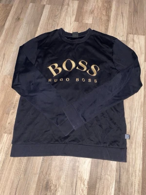 Svart sweatshirt från Hugo Boss - Svart långärmad sweatshirt från Hugo Boss med stort guldfärgat BOSS-tryck på bröstet. Tröjan har rund hals, ribbade muddar vid ärmslut och nederkant samt en klassisk, loose passform. Perfekt för dig som gillar stilrena märkesplagg. Ny pris 1299