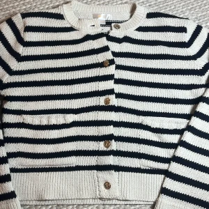 Randig kofta från Gina Tricot - Säljer en snygg stickad kofta från Gina Tricot i storlek 146/152. Koftan är vit med svarta horisontella ränder, har rund hals, långa ärmar och knäpps med fem bruna knappar framtill. Två små fickor framtill. 